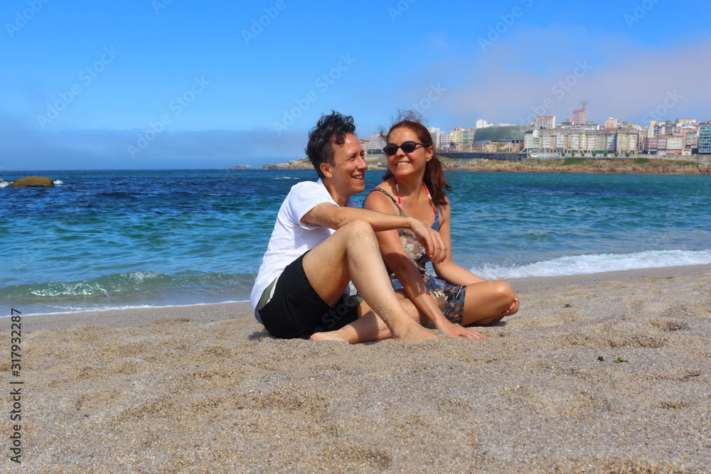 Pareja de un hombre y una mujer sentados en la arena de la playa sonriendo