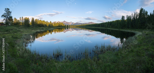 Panorama of a mountain lake overgrown with grass. Snowy mountains, clouds and the moon are reflected in the water. Summer evening, Altai.