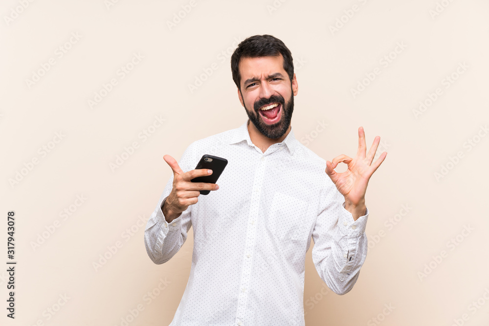 Young man with beard holding a mobile showing ok sign and thumb up gesture Young man with beard holding a mobile showing ok sign and thumb up gesture