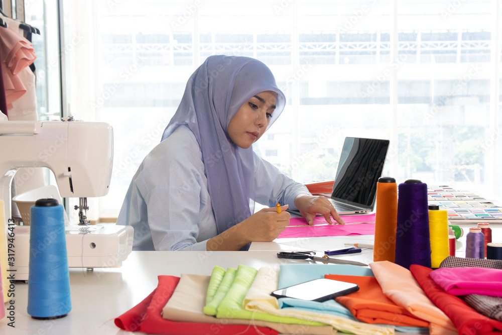 A Muslim woman sitting in the tailoring room for measure the size of the fabric clothing. She is very determined and detailed with the creation of the work. Concept on designing and making product.