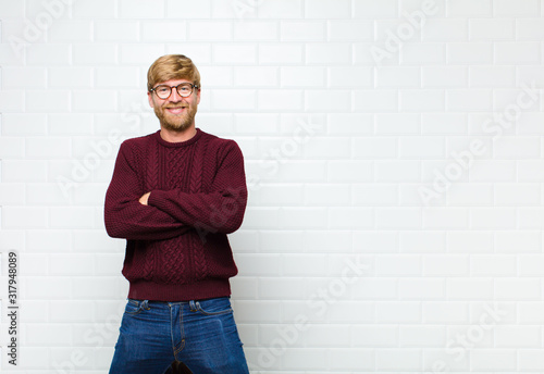 young blonde man looking like a happy, proud and satisfied achiever smiling with arms crossed agaist vintage tiles wall photo