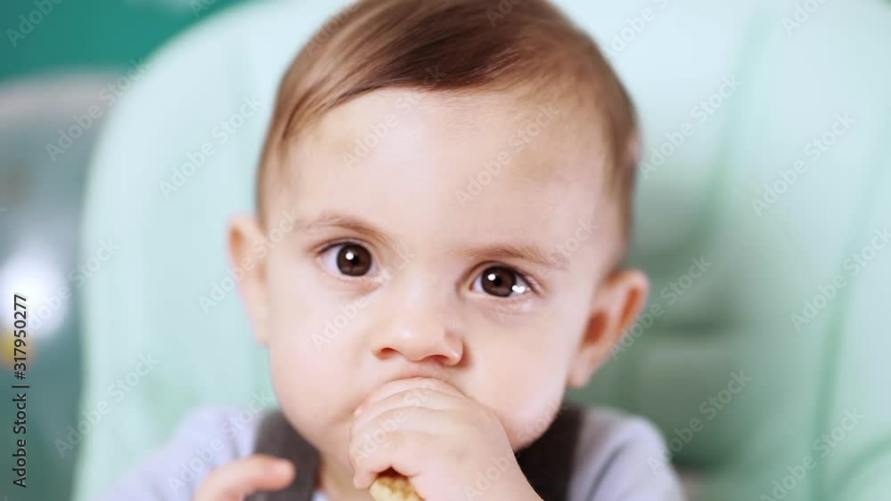 Cute baby boy sitting in his chair and eating delicious cookie. Birthday handsome toddler child with big eyes portrait. Slow motion. Cute baby boy sitting in his chair and eating delicious cookie. Birthday handsome toddler child with big eyes portrait. Slow motion.