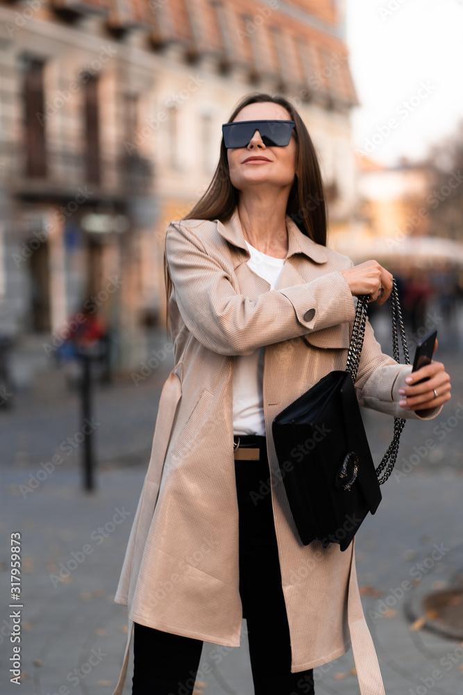 Beautiful brunette woman with phone walking on street.Fashion street style portrait. wearing dark casual trousers, white sweater,creamy coat and sunglasses .Fashion concept.Female business style. Beautiful brunette woman with phone walking on street.Fashion street style portrait. wearing dark casual trousers, white sweater,creamy coat and sunglasses .Fashion concept.Female business style.
