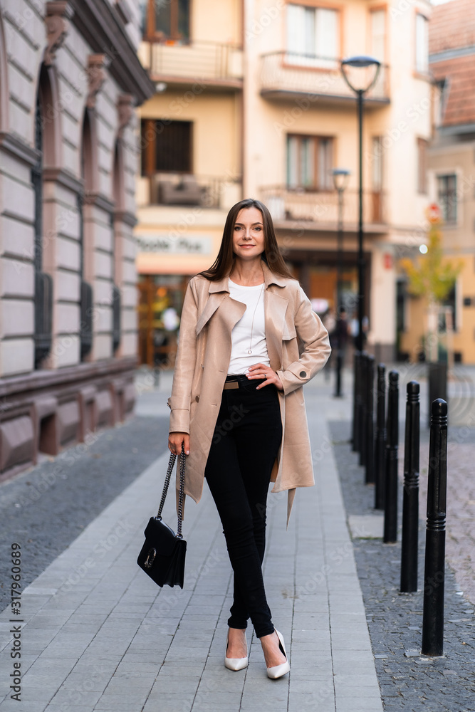 Outdoor photo of brunette lady walking on street background in autumn day.Fashion street style portrait. wearing dark casual trousers, white sweater and creamy coat.Fashion concept.