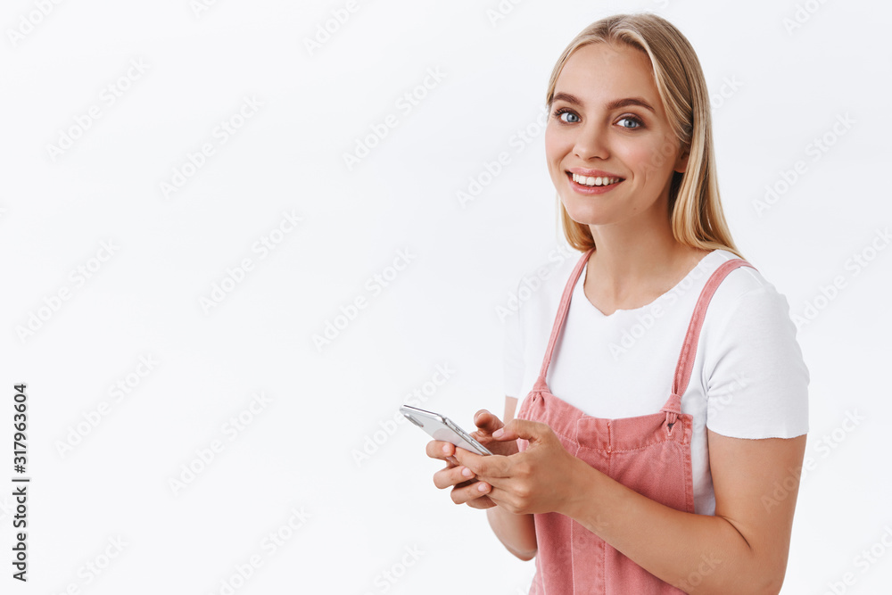 Cheerful young blond female in pink overalls, t-shirt standing half-turned and looking camera with happy smile, holding smartphone, messaging or using social media when someone called her name Cheerful young blond female in pink overalls, t-shirt standing half-turned and looking camera with happy smile, holding smartphone, messaging or using social media when someone called her name