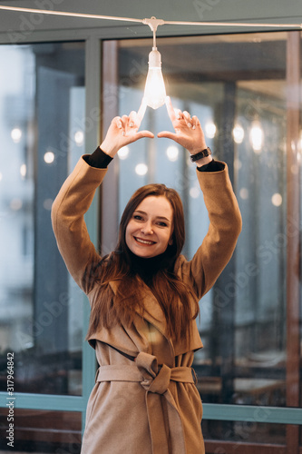 Outdoor fashion portrait of stylish young woman having fun with lamp , laughing, Urban city street style wearing dark casual trousers, white sweater,creamy coat. and sunglasses Fashion concept.