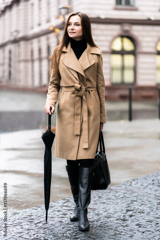 Outdoor photo of brunette lady posing with black umbrella in rainy autumn day.Fashion street style portrait. wearing dark casual trousers, white sweater and creamy coat.Fashion concept. Outdoor photo of brunette lady posing with black umbrella in rainy autumn day.Fashion street style portrait. wearing dark casual trousers, white sweater and creamy coat.Fashion concept.