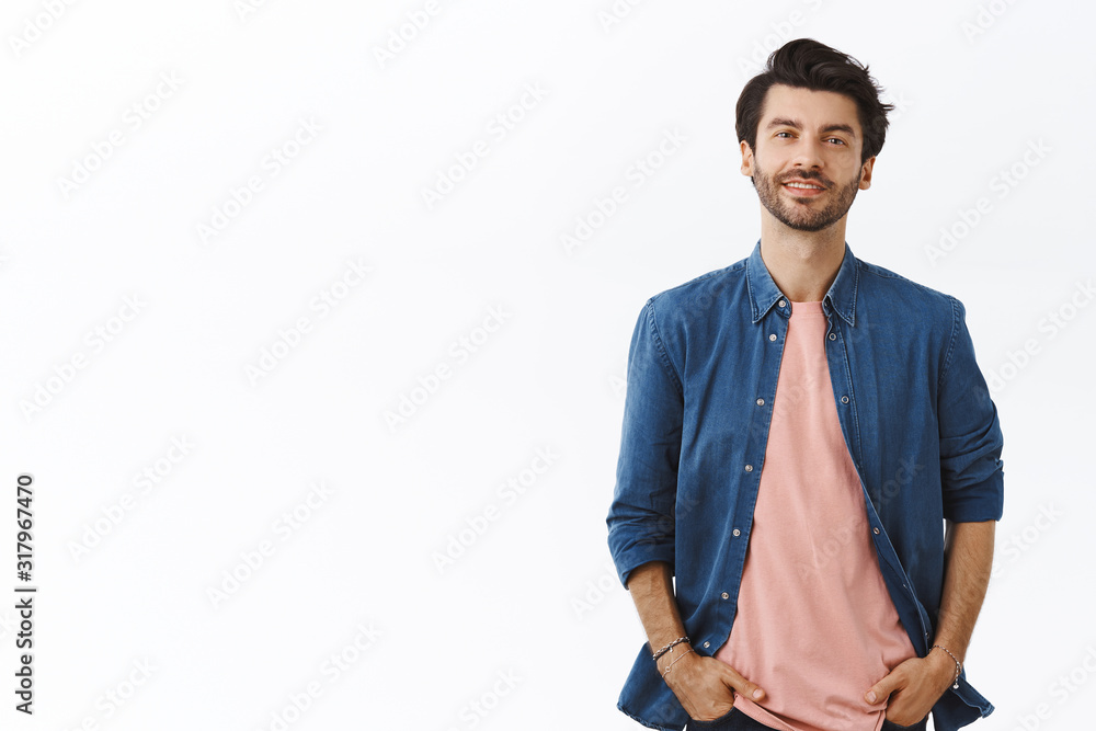 Cheerful, bearded handsome young man with messy hairstyle, standing relaxed in casual pose with hands in pockets, smiling, ordinary guy shopping in mall, buy gifts. Male look camera enthusiastic - obrazy, fototapety, plakaty 