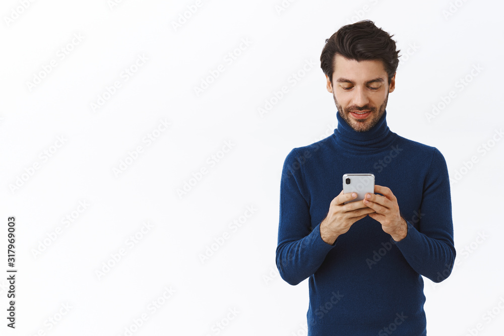 Handsome bearded stylish businessman in trendy high neck sweater, holding smartphone, taking notes in mobile phone, looking at device as messaging, checking events going nearby, white background Handsome bearded stylish businessman in trendy high neck sweater, holding smartphone, taking notes in mobile phone, looking at device as messaging, checking events going nearby, white background