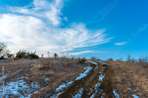 Dirt road leading to the top of the hill at wintertime, vibrant blue sky with white clouds.