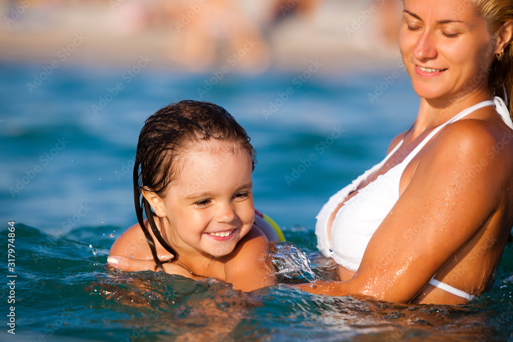 Young blond mother in blue bikini standing in water and helping to swim her small smiling daughter in sunshine on summer day. Family vacations and travelling concept Young blond mother in blue bikini standing in water and helping to swim her small smiling daughter in sunshine on summer day. Family vacations and travelling concept