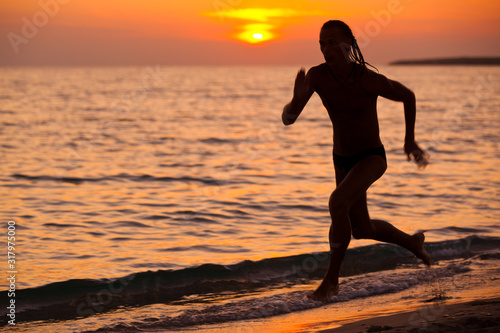 Silhouette of young athletic man running on sea water edge during colorful golden sunset on summer day. Vacations  travelling  active healthy lifestyle concept