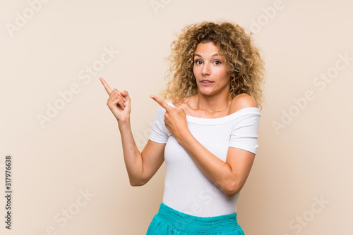 Young blonde woman with curly hair over isolated background frightened and pointing to the side © luismolinero