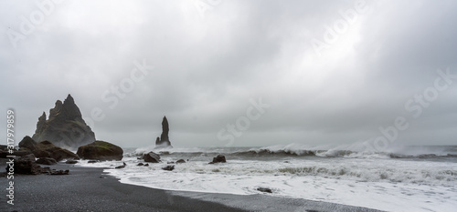 Panorama of Waves crashing in on shore on Reynisfjara during a storm in Iceland. Basalt sculpture columns raises up from the sea in the background. Black lava sand infront. 