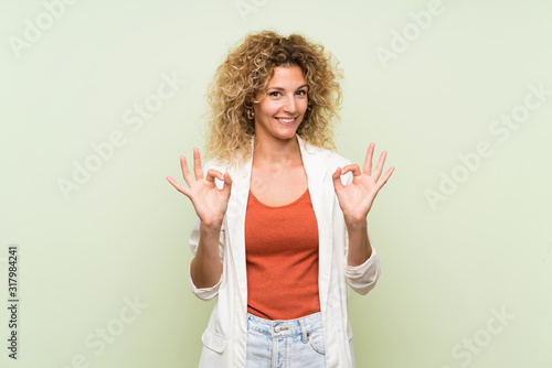Young blonde woman with curly hair over isolated green background showing an ok sign with fingers
