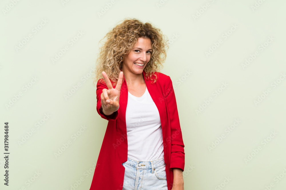 Young blonde woman with curly hair over isolated green background smiling and showing victory sign Young blonde woman with curly hair over isolated green background smiling and showing victory sign