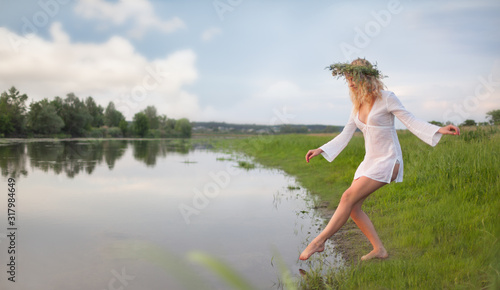Young sexy beautiful blond woman in white mini dress and floral wreath standing and trying water on summer day
