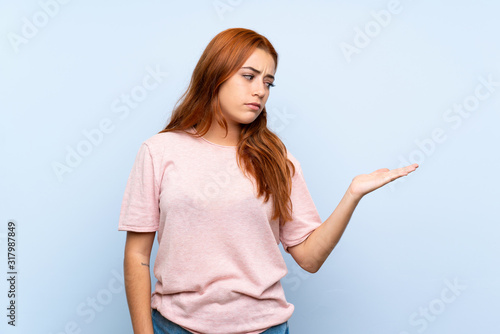 Teenager redhead girl over isolated blue background holding copyspace with doubts