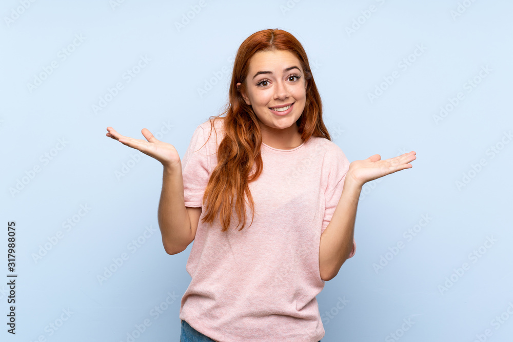 Teenager redhead girl over isolated blue background making doubts gesture