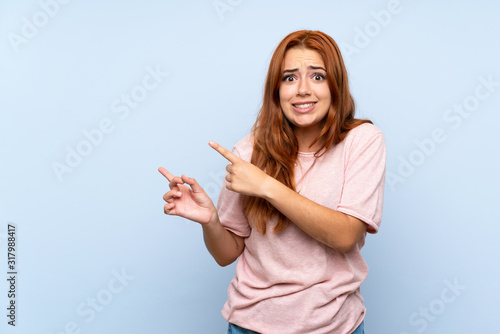 Teenager redhead girl over isolated blue background frightened and pointing to the side