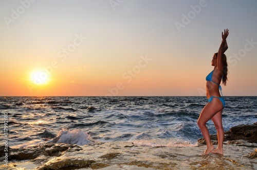Young woman in bikini standing on rocks and enjoying drops of waves and sunset