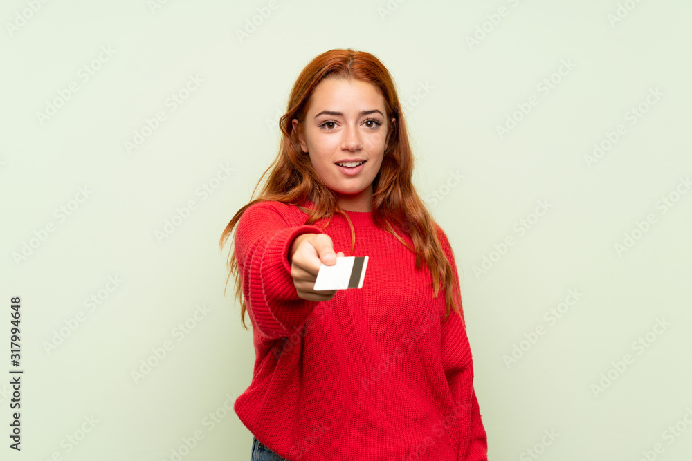 Teenager redhead girl with sweater over isolated green background holding a credit card Teenager redhead girl with sweater over isolated green background holding a credit card