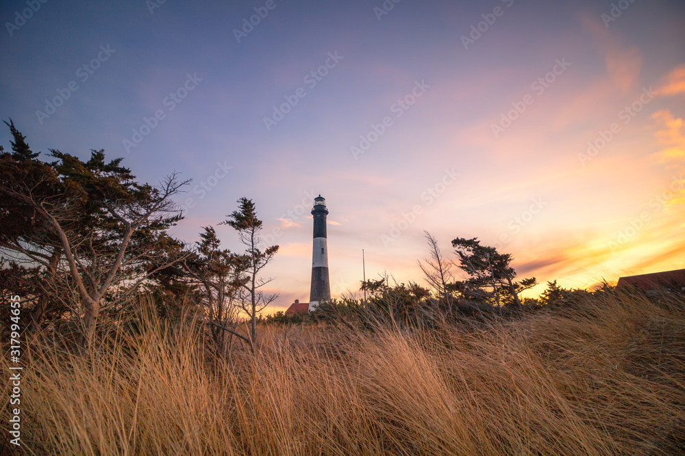 Golden sunset light illuminating beach grass in front of a stone lighthouse, with wispy clouds overhead in the sky. Fire Island National Seashore, New York. Golden sunset light illuminating beach grass in front of a stone lighthouse, with wispy clouds overhead in the sky. Fire Island National Seashore, New York.