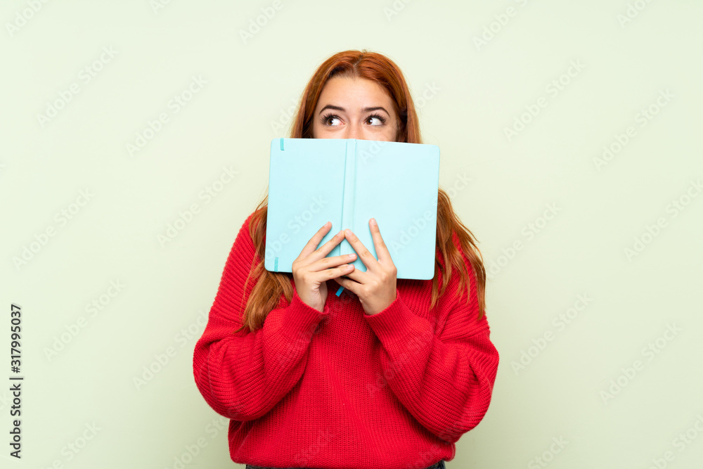 Teenager redhead girl with sweater over isolated green background holding and reading a book Teenager redhead girl with sweater over isolated green background holding and reading a book