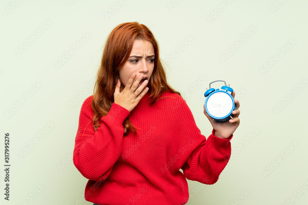 Teenager redhead girl with sweater over isolated green background holding vintage alarm clock Teenager redhead girl with sweater over isolated green background holding vintage alarm clock