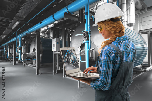  Attractive Caucasoid engineer woman with laptop inspect modern industrial gas boiler room. Heating gas boilers, pipelines, valves. Mixed media