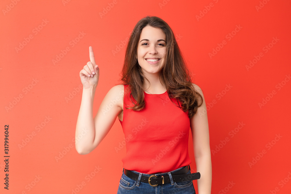 young pretty woman smiling cheerfully and happily, pointing upwards with one hand to copy space against red wall - obrazy, fototapety, plakaty 
