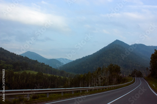 Evening. Asphalt road in the mountains with a dividing strip. Fencing and trees along the road. Mountains, hills