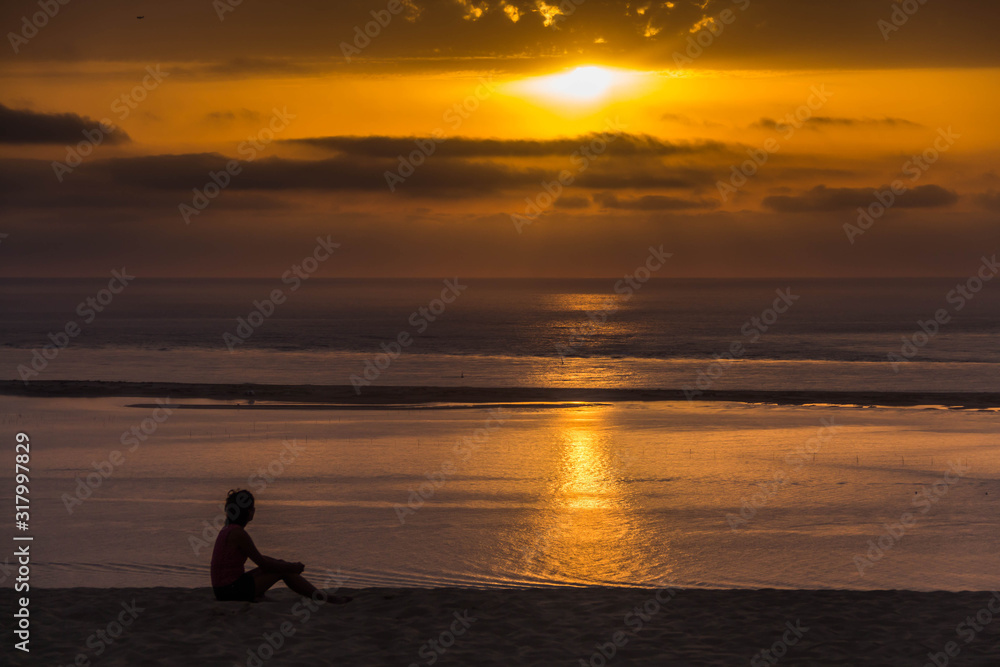 Side view of a young woman seating on the top of the Grande Dune du Pilat and seeing on the Atlantic Ocean and yellow sunset
