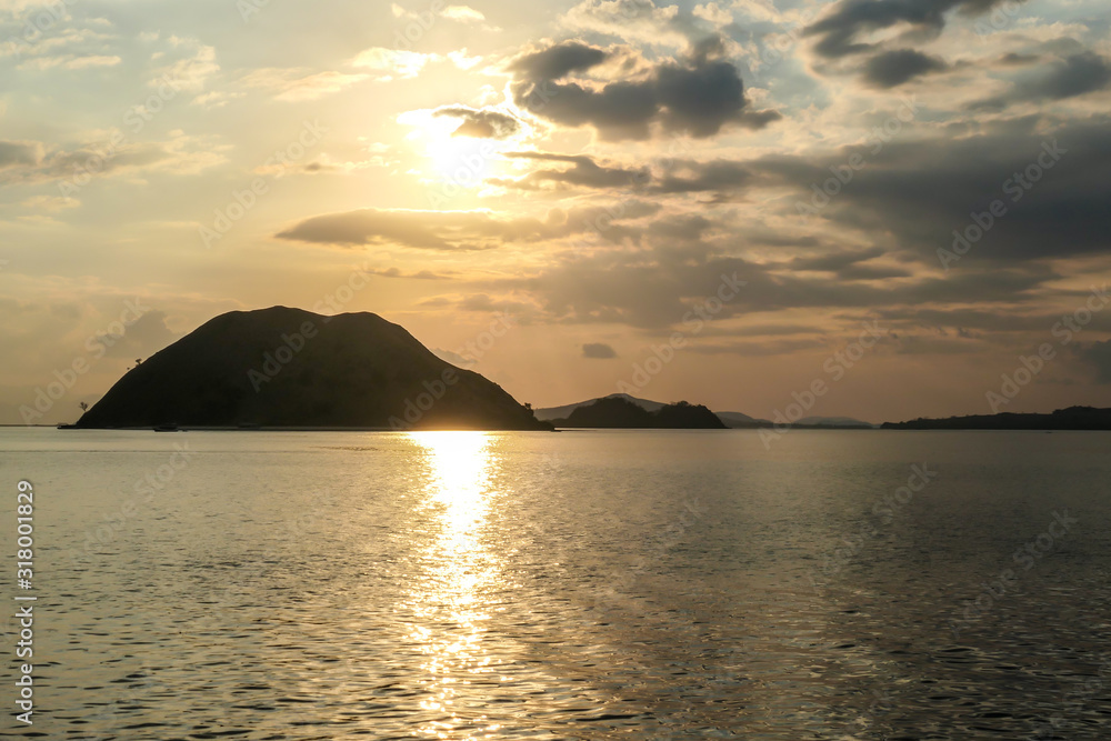 Golden hour in Komodo National Park, Indonesia. Sun starts to set behind one of the islands, leaving a golden path on the calm surface of the sea. Golden hour in Komodo National Park, Indonesia. Sun starts to set behind one of the islands, leaving a golden path on the calm surface of the sea.