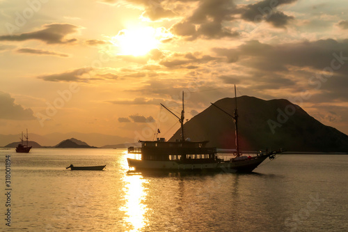 Fototapeta Naklejka Na Ścianę i Meble -  Golden hour in Komodo National Park, Indonesia. Sun sets behind one of the islands, leaving a golden path on the calm surface of sea. Some boats anchored to the shore, enjoying the natural spectacle.