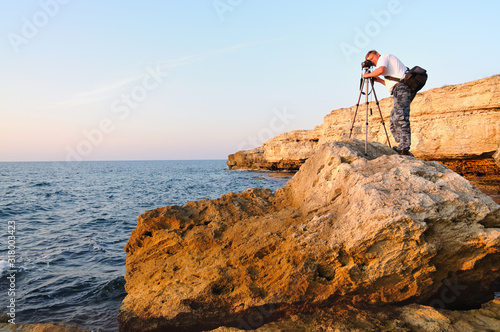 Man standing on rock and taking photo with tripod over Black sea coastline with clear blue waters and blue sky in Crimea. Natural background and wallpaper