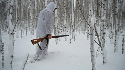 A hunter in the winter in the woods in a white camouflage suit hunts a hare.