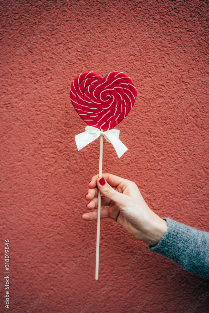 Big caramel heart on a stick in an elegant female hand on a pink background. Vertical image. Valentine's day concept. Big caramel heart on a stick in an elegant female hand on a pink background. Vertical image. Valentine's day concept.