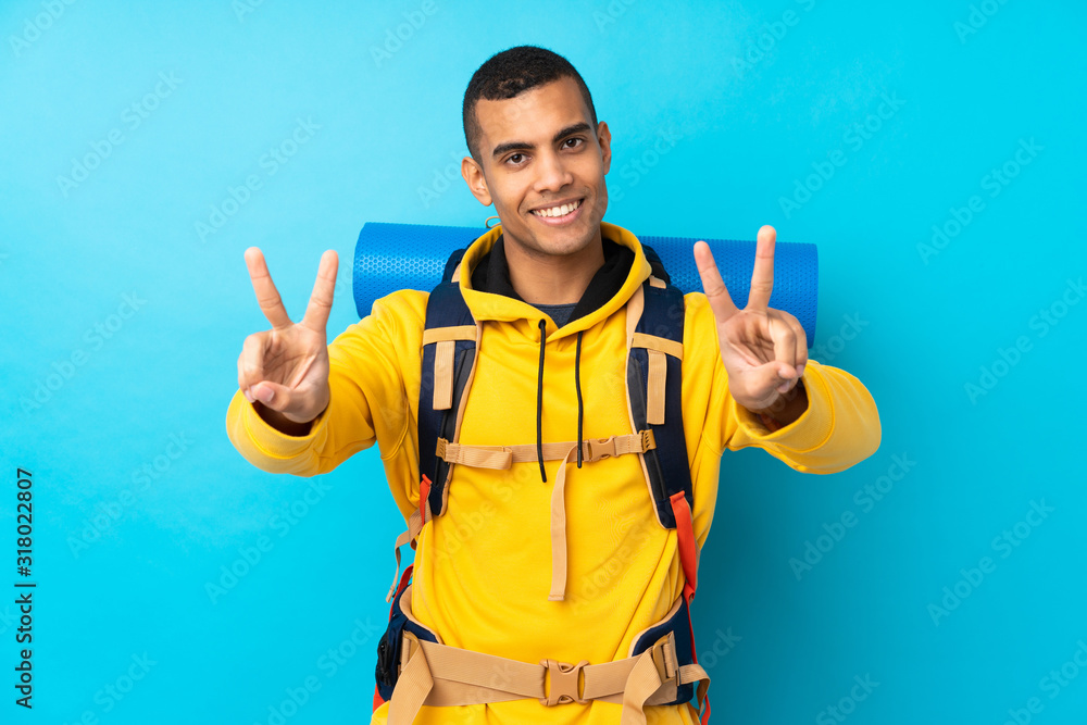 Young mountaineer man with a big backpack over isolated blue background smiling and showing victory sign Young mountaineer man with a big backpack over isolated blue background smiling and showing victory sign
