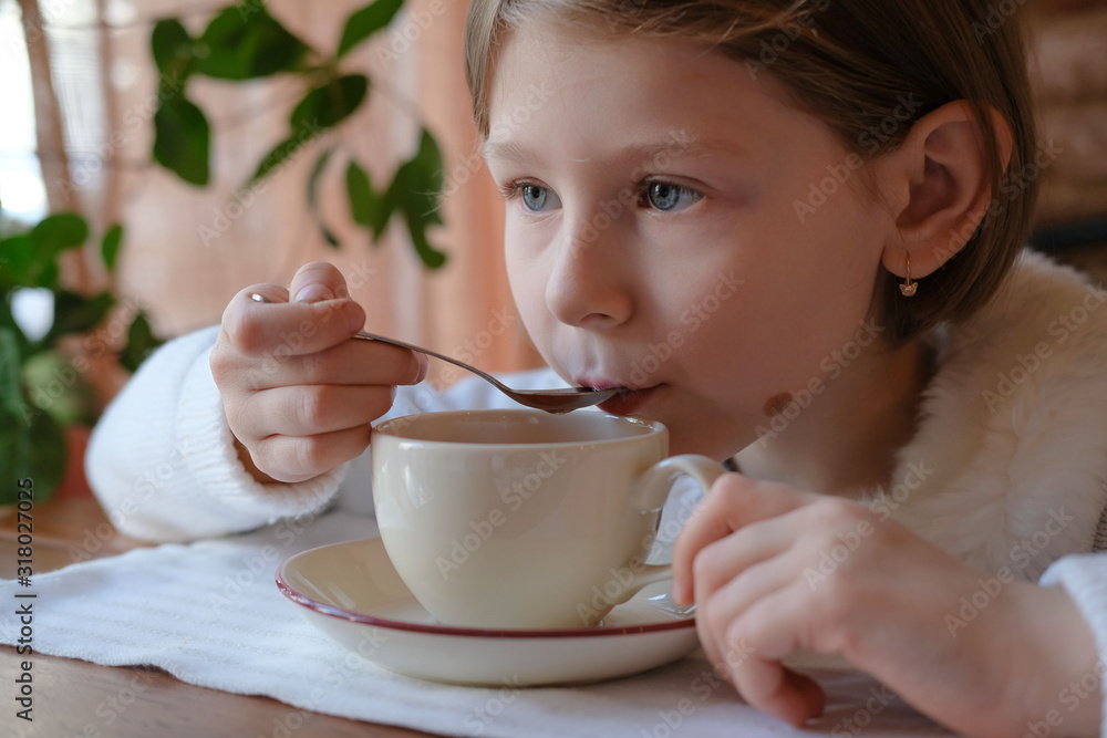 a little girl in a white sweater sits in a cafe by the window and drinks hot drink tea, coffee, cocoa from a spoon. on the table in front of the girl is a cup a little girl in a white sweater sits in a cafe by the window and drinks hot drink tea, coffee, cocoa from a spoon. on the table in front of the girl is a cup