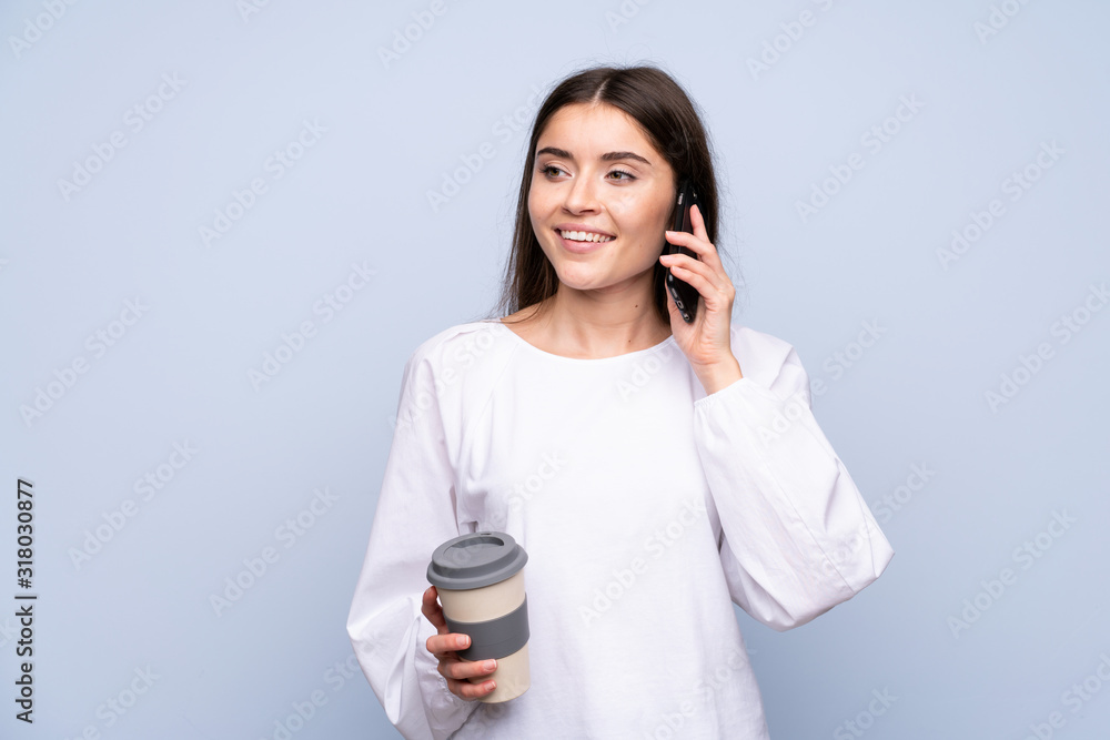 Young woman over isolated blue background holding coffee to take away and a mobile Young woman over isolated blue background holding coffee to take away and a mobile