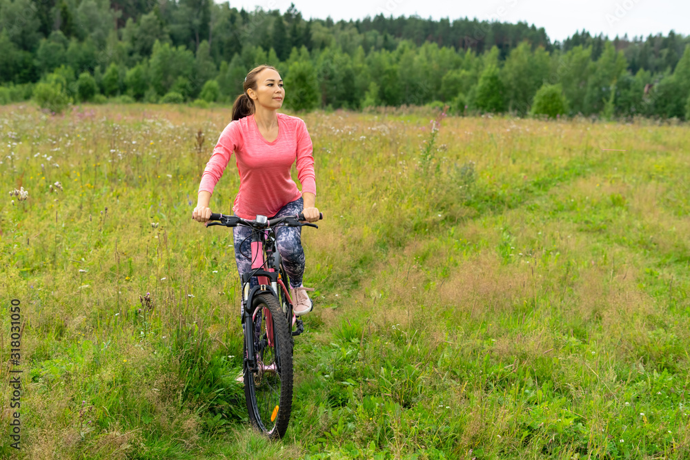Asian woman cycling pink bike. Happy fitness girl is riding a bicycle across the green field at summer day. Smiling attractive sporty girl recreation on bicycle outdoors. Fitness Travel Workout.
