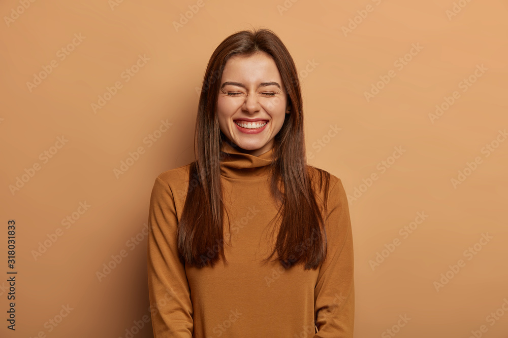 Φωτογραφία Pretty amused woman has long dark straight hair, laughs from joy, wears brown neck sweater, smirks face with pleasure, closes eyes, stands against beige bakground