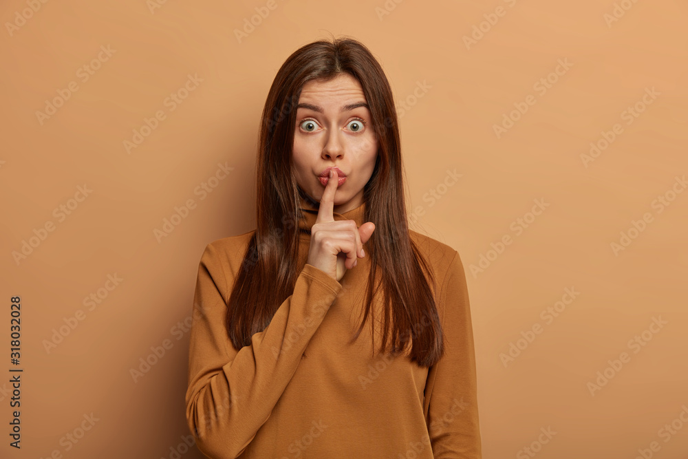 Φωτογραφία Brunette European woman keeps index finger on lips, make hush gesture, keeps secret, shocked by rumors, has healthy skin, wears casual brown jumper, poses indoor, asks to sit quiet