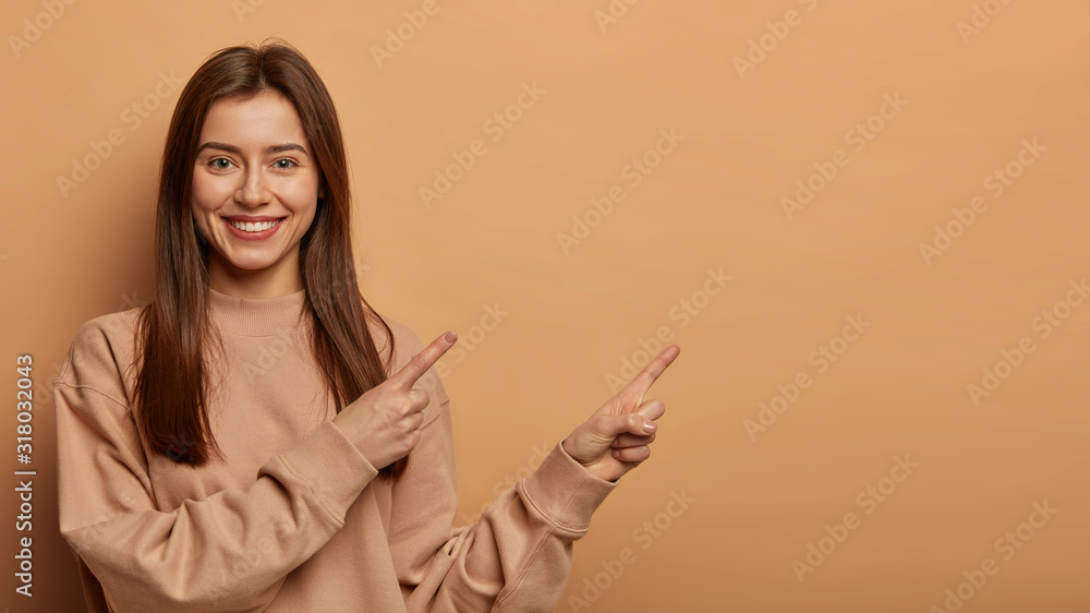 Look, advertise here! Dark haired pretty woman with appealing smile gives positive attitude towards good deal, expresses her recommendation while pointing on right side, wears brown sweater. Look, advertise here! Dark haired pretty woman with appealing smile gives positive attitude towards good deal, expresses her recommendation while pointing on right side, wears brown sweater.