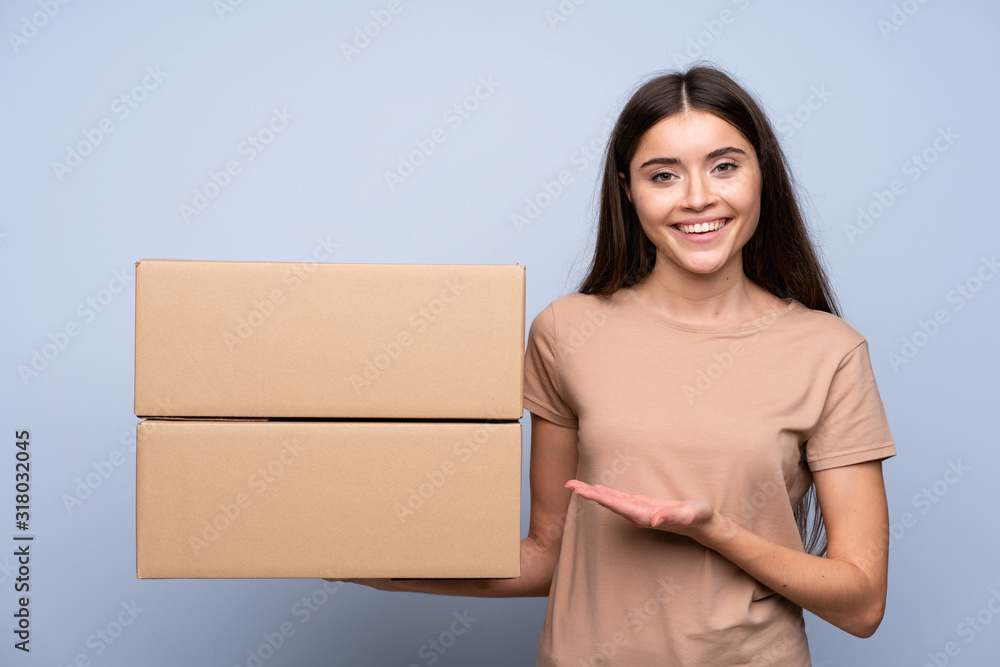Young woman over isolated blue background holding a box to move it to another site Young woman over isolated blue background holding a box to move it to another site