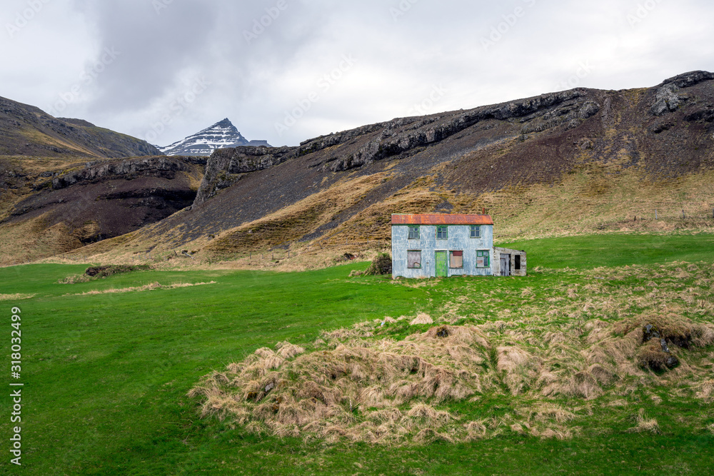 Bulandstindur mountain peak with snow on it. Blue abandoned farmhouse infront of it. Near Djupivogur in Iceland next to the ring road furing road trip. Icelandic landscape concept.