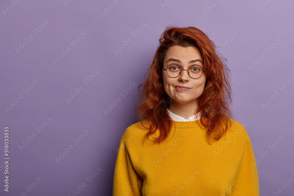 Φωτογραφία Lovely redhead wonan purses lips, has serious face expression, healthy skin, wears optical glasses, yellow casual jumper, isolated over purple background, copy space