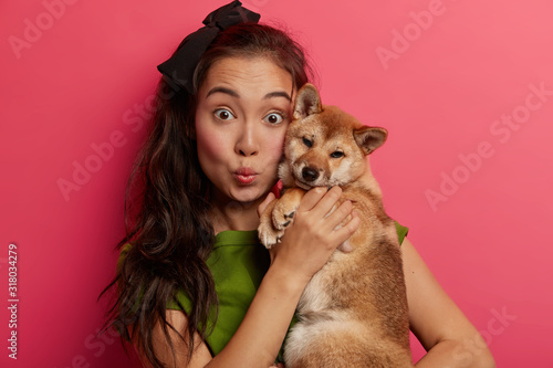 Close up shot of surprised dark haired ethnic woman holds Shiba Inu dog near face, keeps lips rounded, feels closeness with loyal animal, grows pet with love and care, isolated on pink background