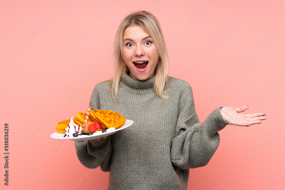 Young blonde woman holding waffles over isolated pink background with shocked facial expression Young blonde woman holding waffles over isolated pink background with shocked facial expression